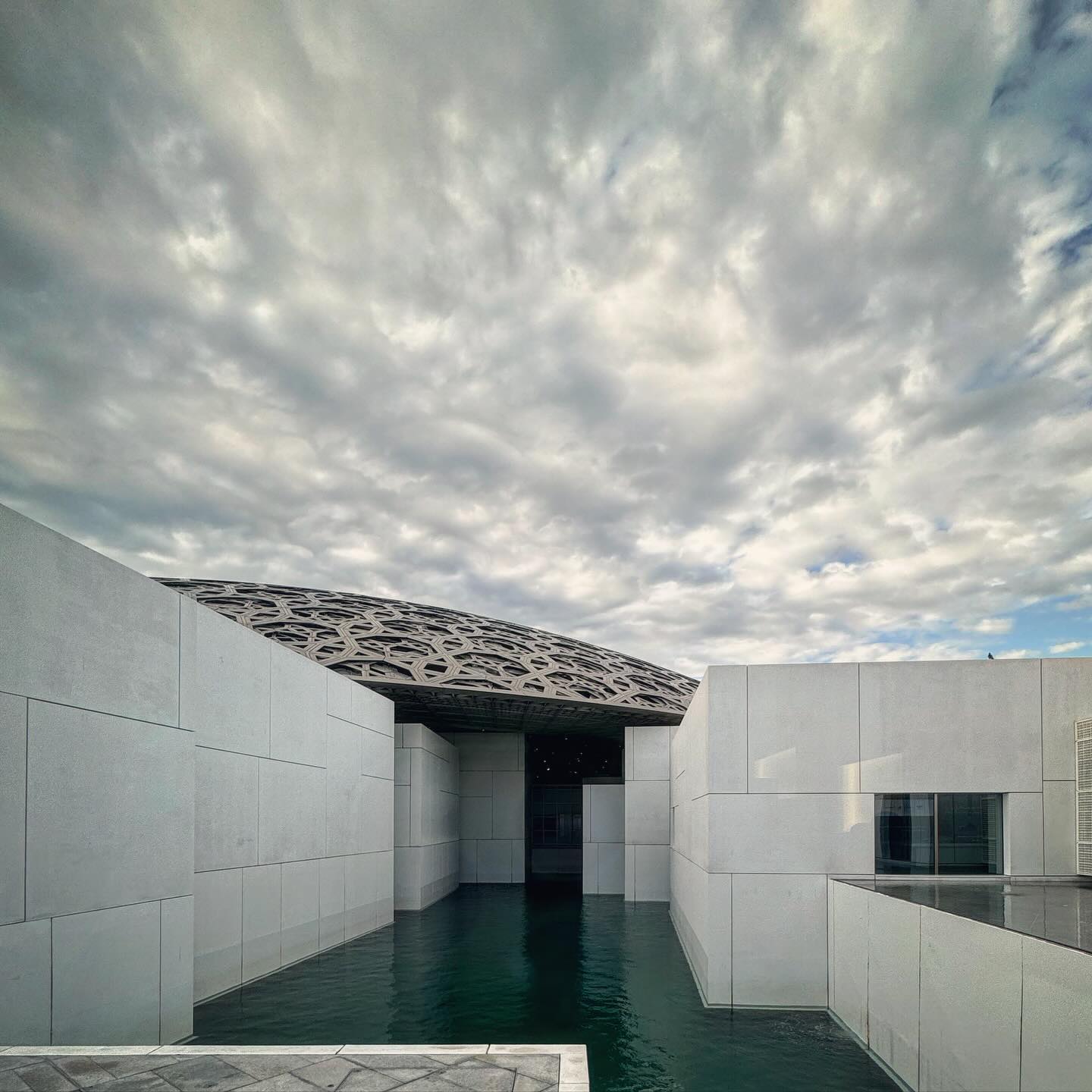 Low pale museum walls frame a pool beneath a clouded sky.