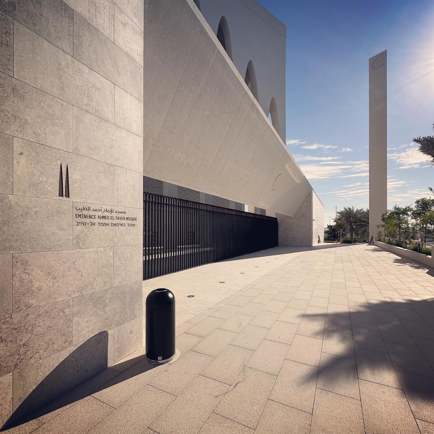 A pale stone wall and slender vertical forms beside a shaded pedestrian route.