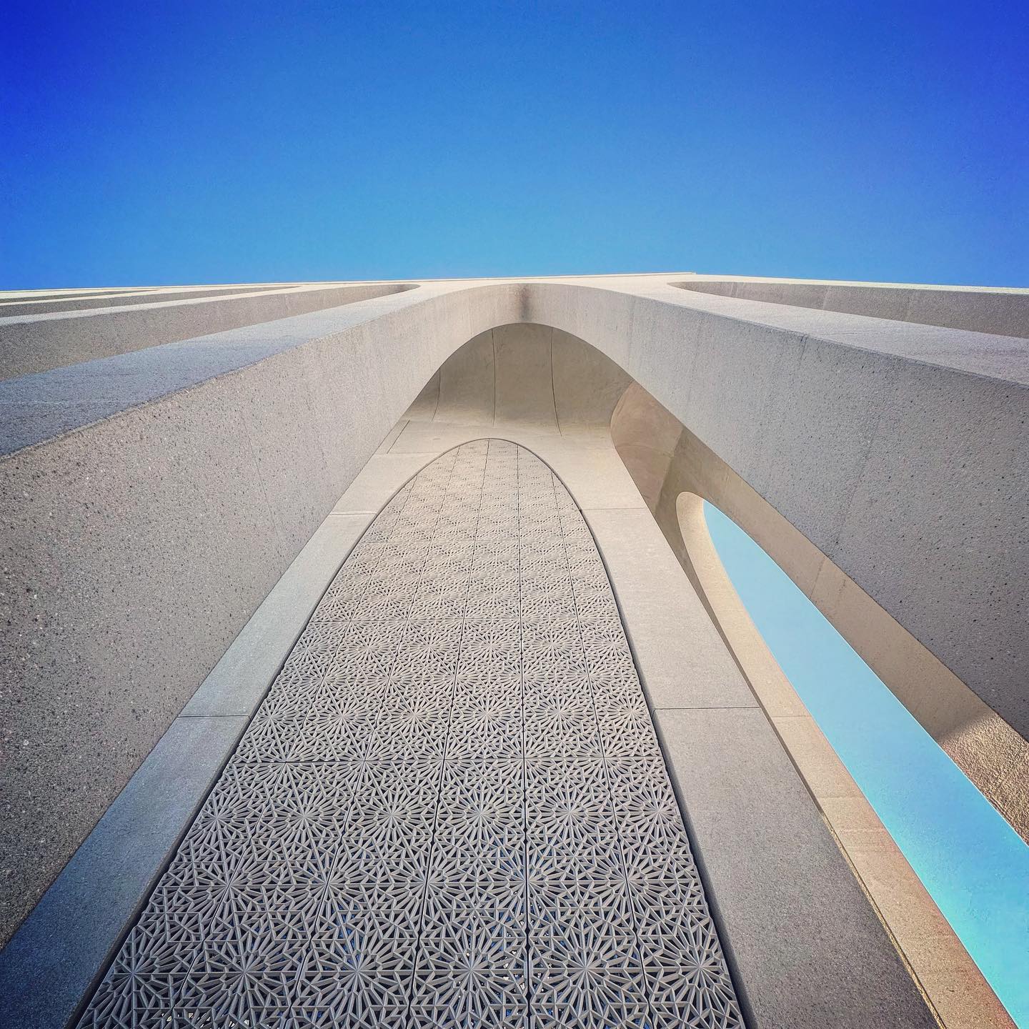 An upward view of pale curved stonework and a geometric screen against a clear blue sky.