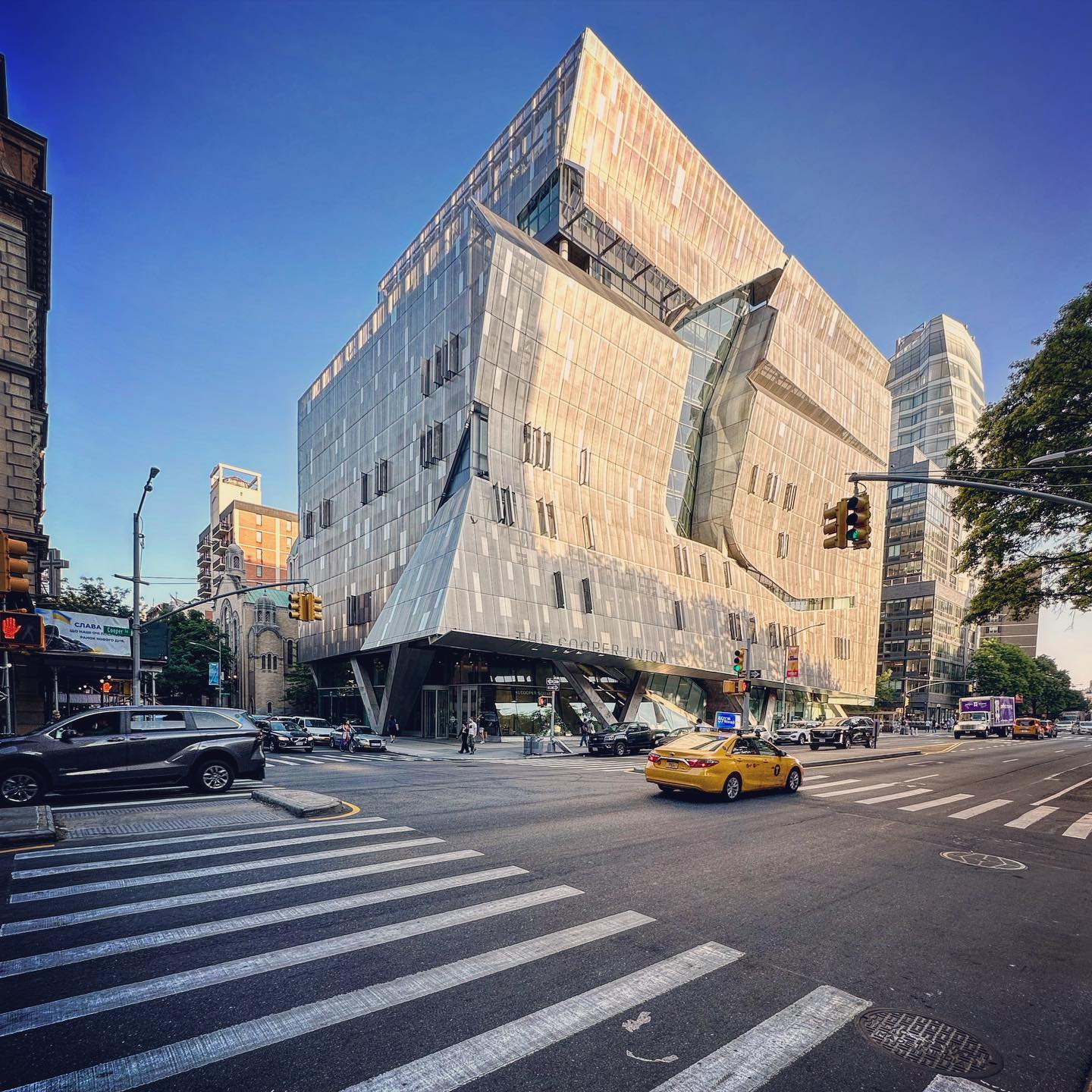An angled residential building rises above a street crossing and yellow taxi.