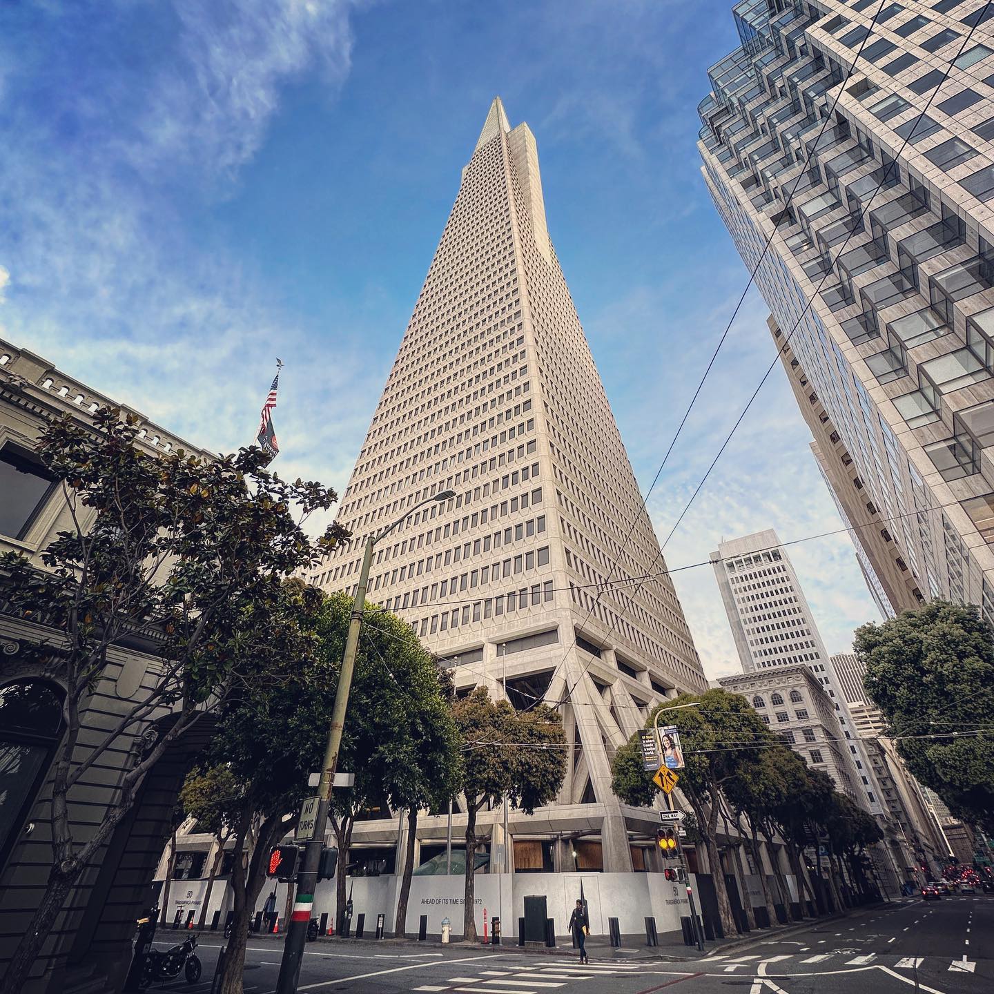 The pointed tower rises above street-level planting between neighbouring buildings.