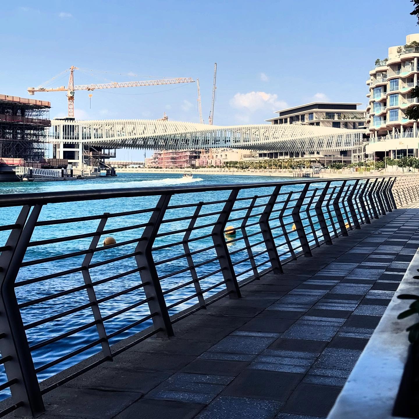 A pedestrian promenade and dark railings follow a blue canal towards a bridge and waterfront buildings.