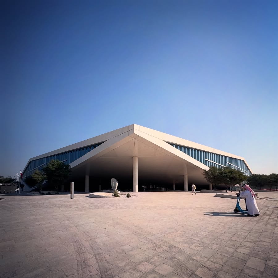 An angular pale roof extends over a broad shaded entrance and open forecourt.