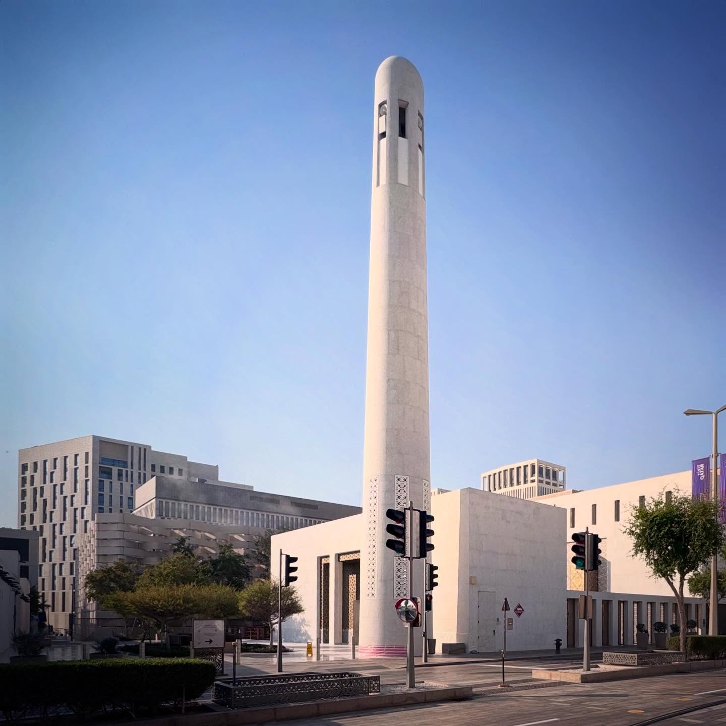 A slender pale minaret rises above low buildings and a planted street edge.