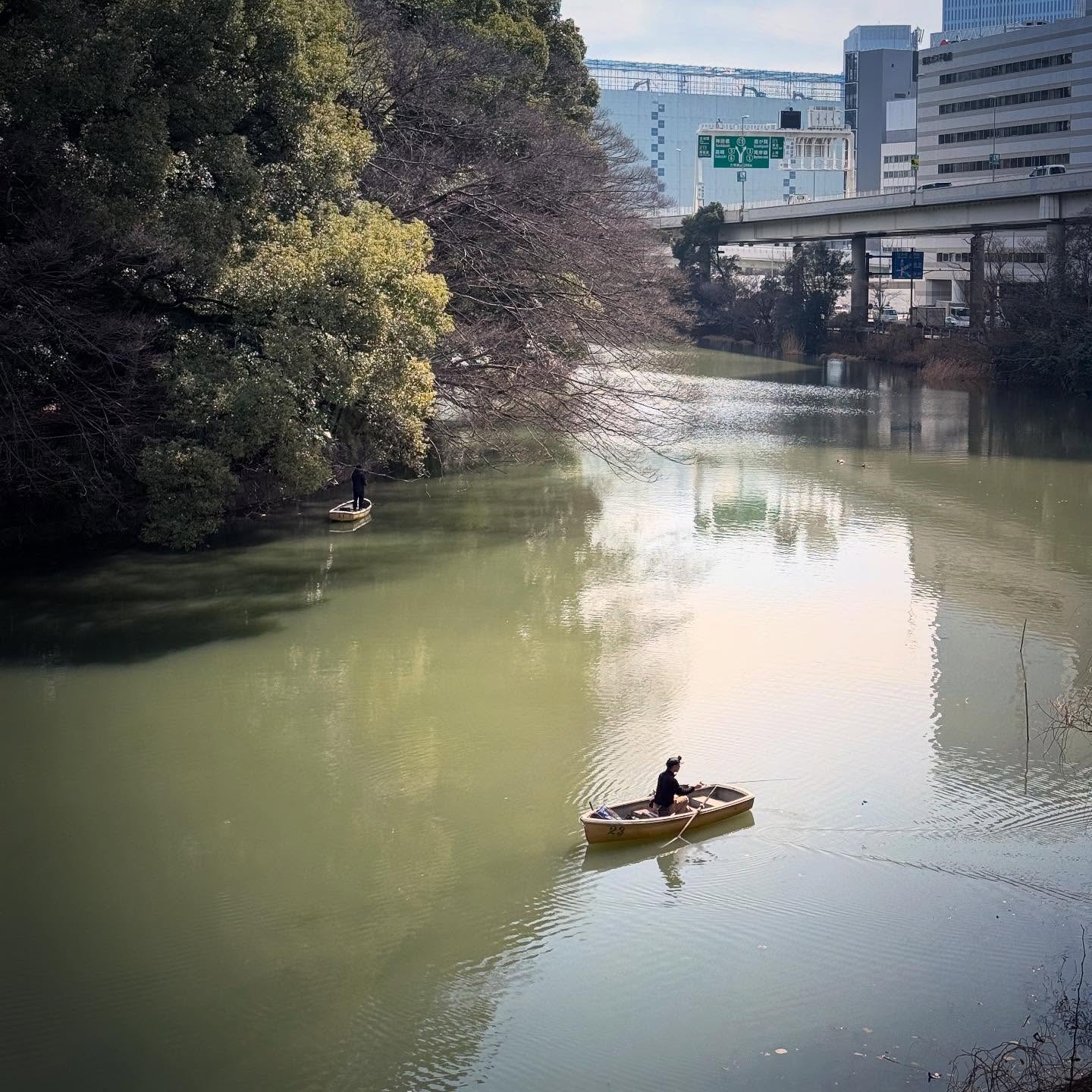 A small boat on a quiet stretch of water, framed by trees and Tokyo buildings.