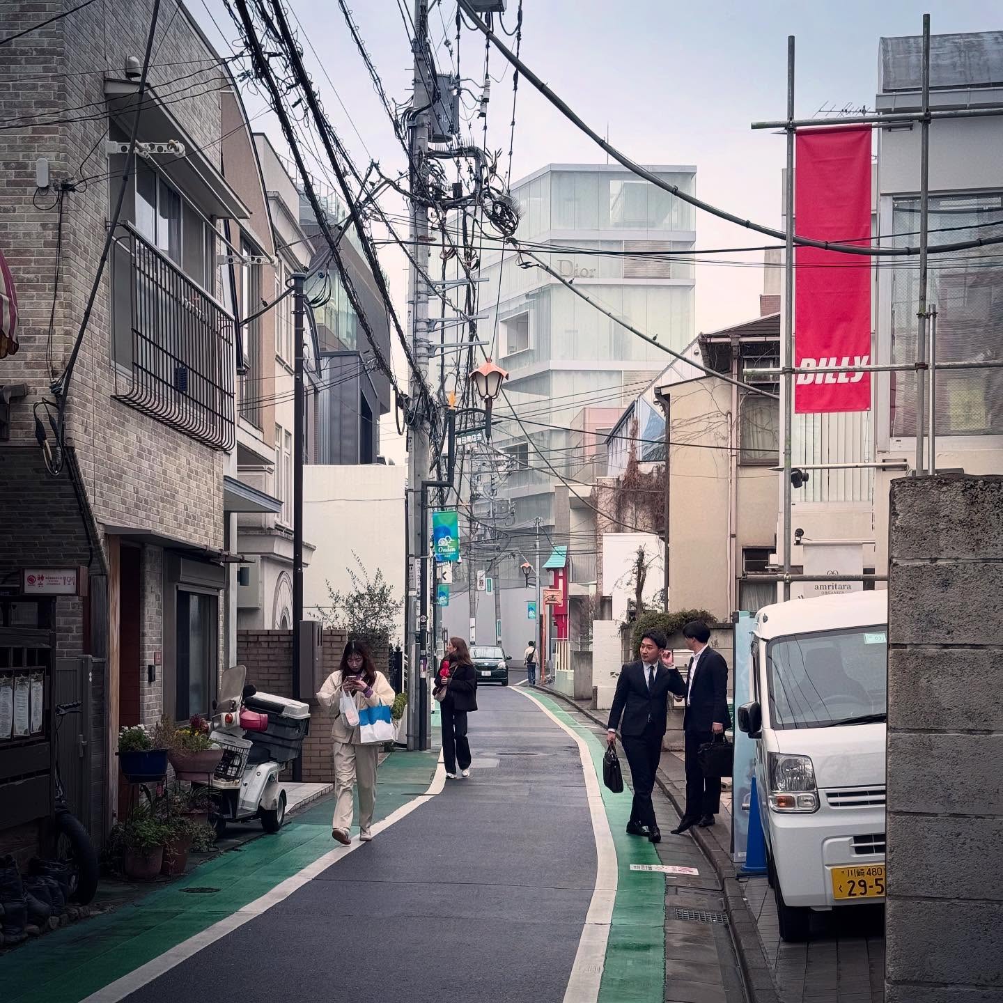 A narrow Tokyo lane with small shops, pedestrians and overhead cables.
