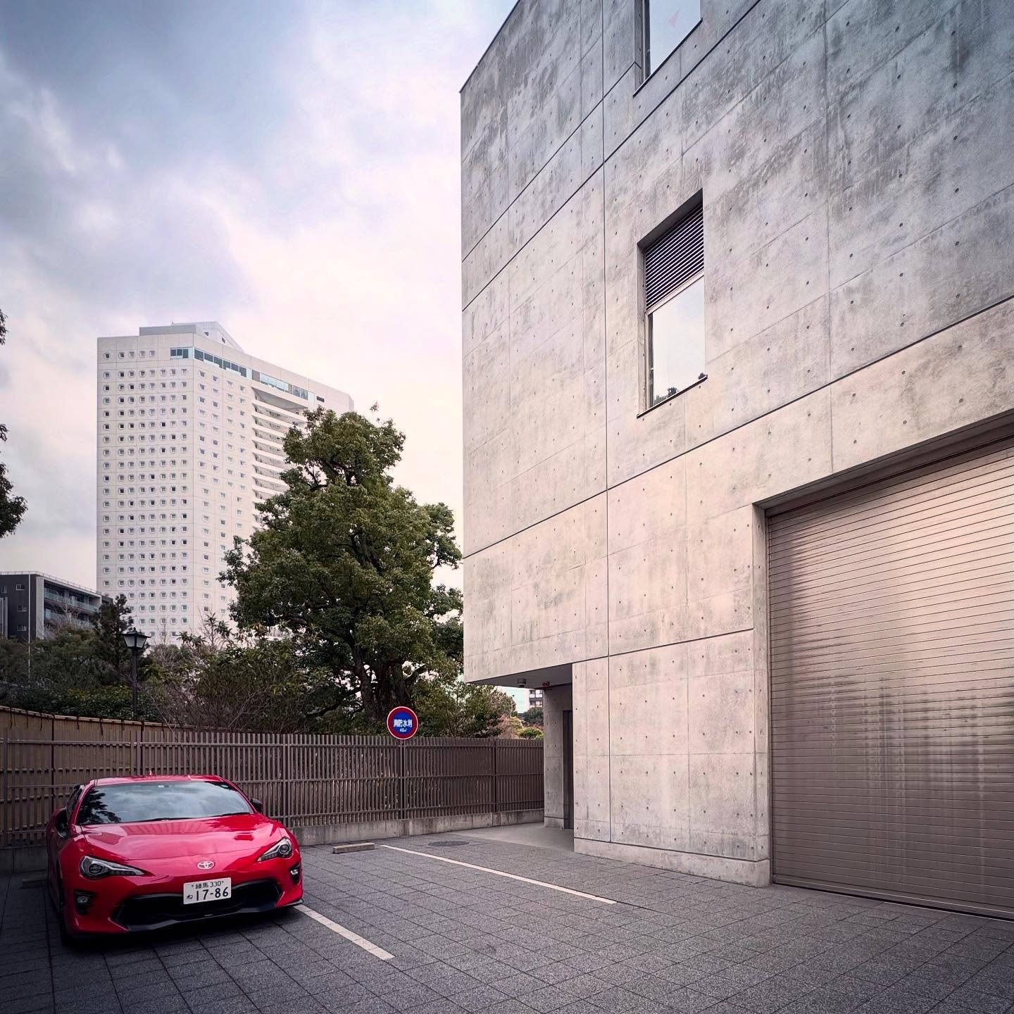 A red car beside a raw concrete façade on a Tokyo street.