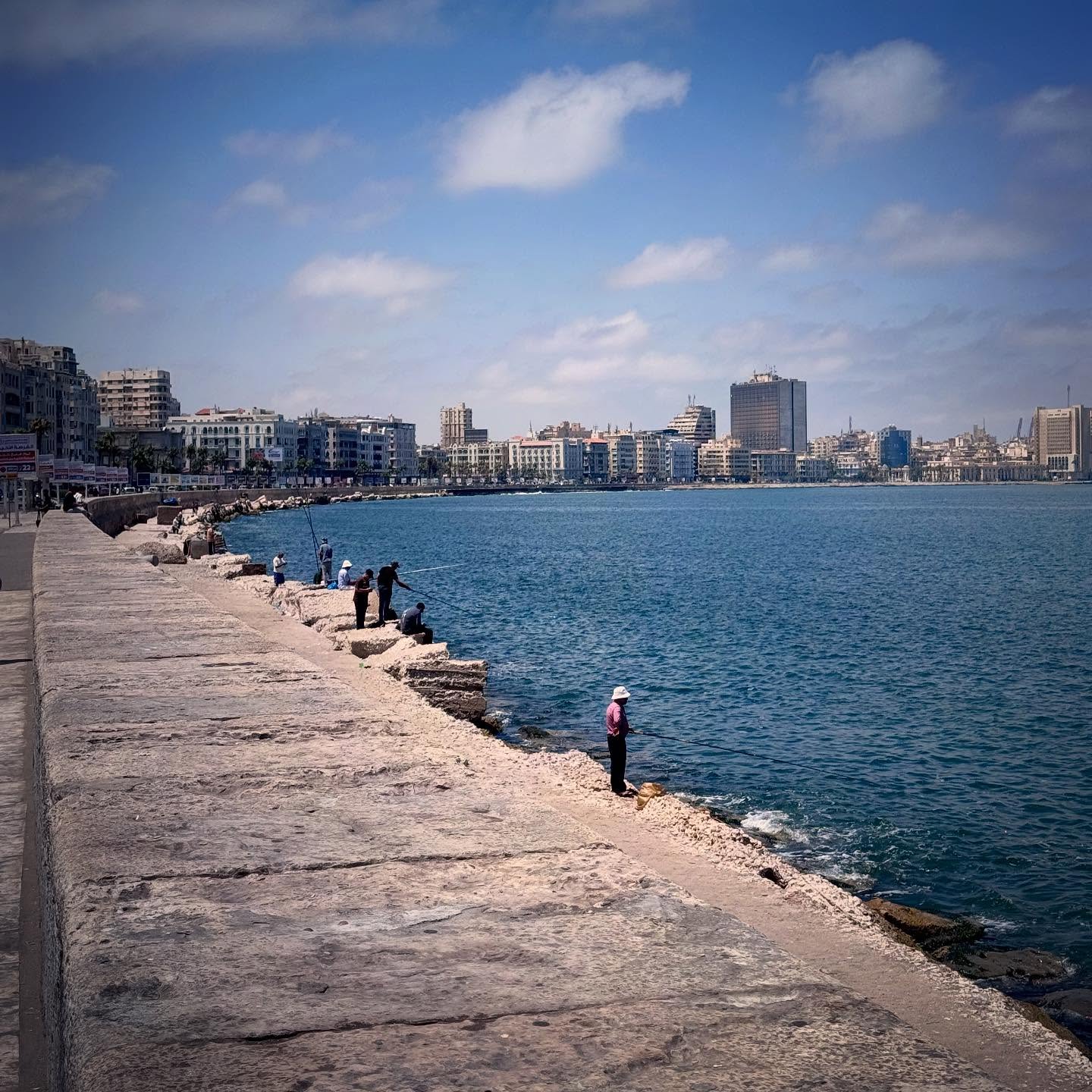 The stone waterfront curves beside blue water towards the city skyline.