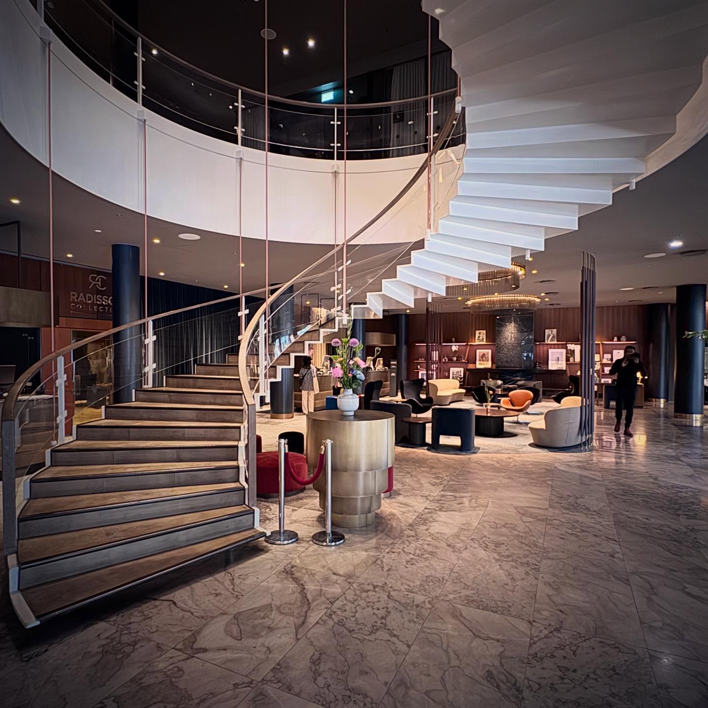 A sweeping suspended stair curves above the hotel lobby and seating areas.