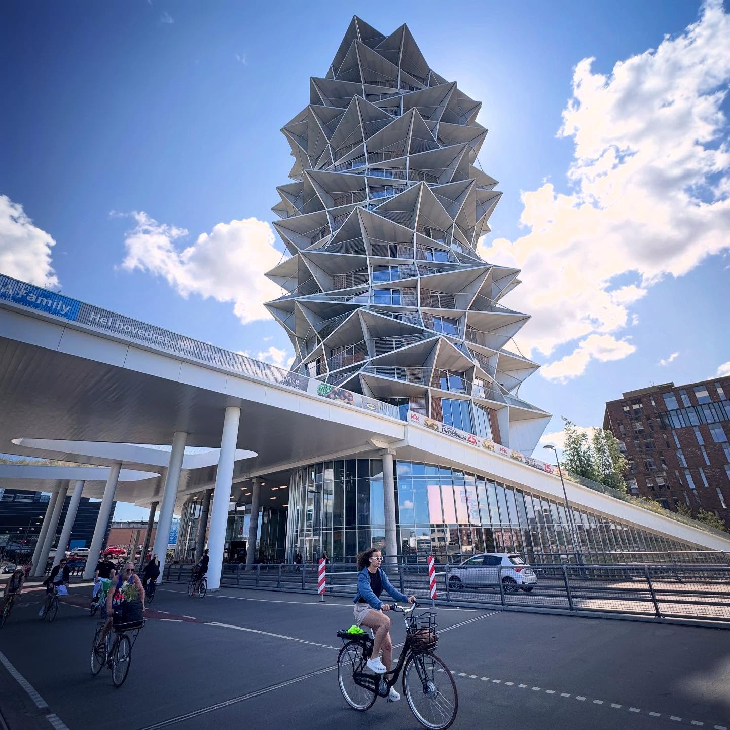 A residential tower with projecting angular balconies above cyclists and a glazed podium.