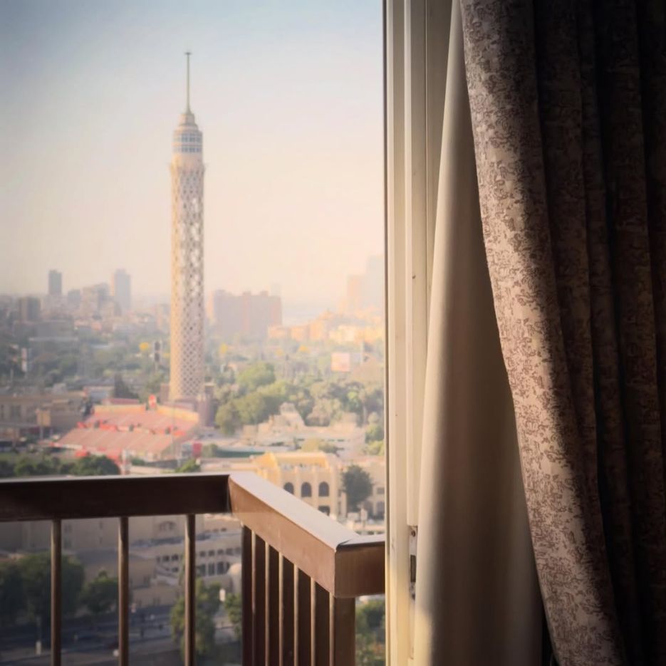 Cairo Tower seen through a balcony doorway, framed by curtains and a balustrade.