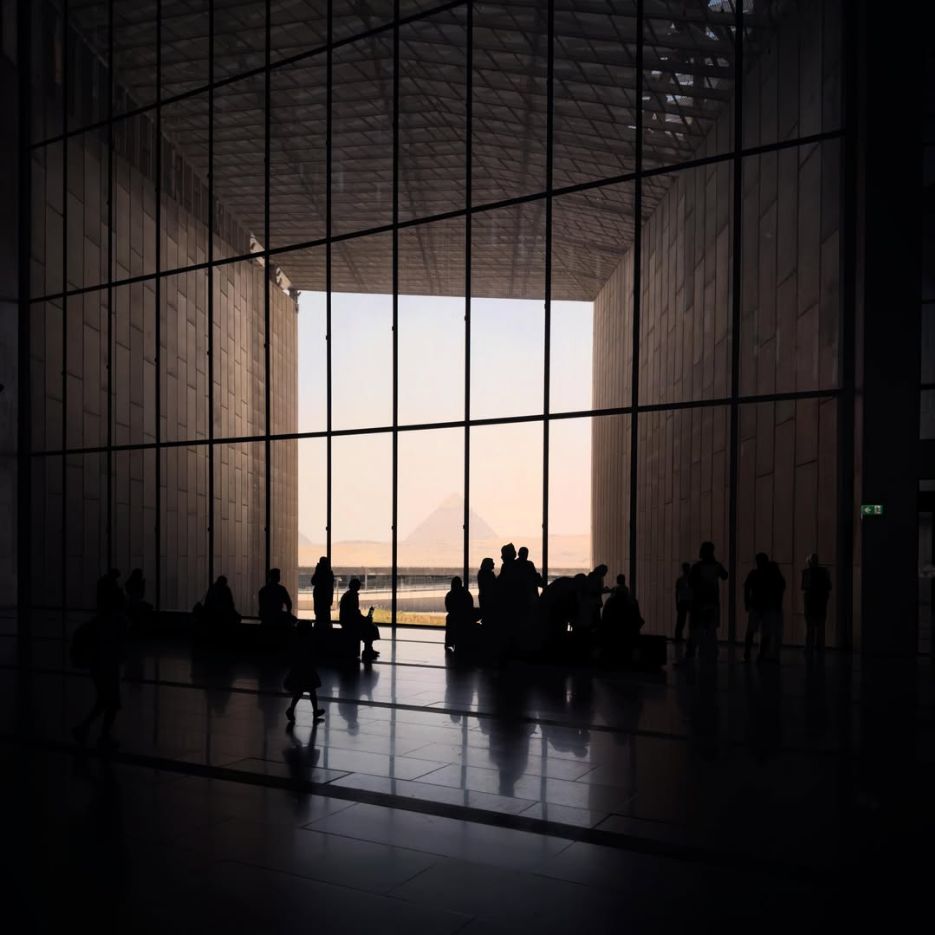 Visitors silhouetted against the glazed hall of the Grand Egyptian Museum, with a pyramid in the distance.